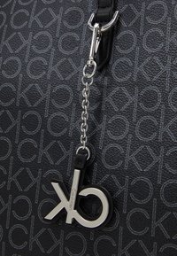 Sac à main noir à texture, présentant un motif en relief du logo, des accessoires en métal argenté et un porte-clés avec un accent en logo circulaire.