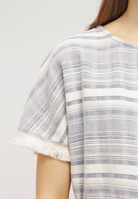 Raidallinen pusero vaaleanharmaana ja valkoisena, jossa on pehmeä kangas, laatikkomainen muoto, fringed-helma ja teksturoitu kudonta vaakasuorilla viivoilla.