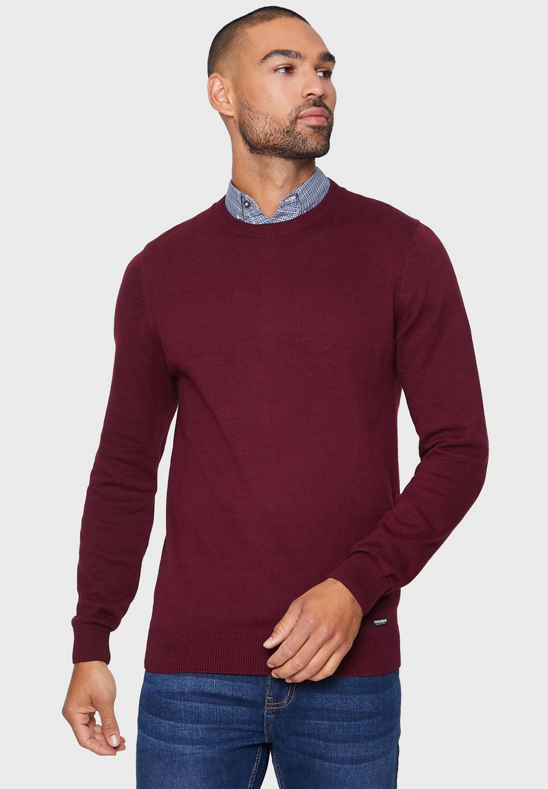 Maglione bordeaux a collo rotondo in tessuto a maglia liscia, con polsini e orlo a costine. Indossato sopra una camicia a quadri blu, evidenziando un look a strati.