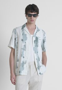 Camicia leggera a maniche corte con base bianca e motivi astratti in teal, dotata di colletto a camicia e chiusura con bottoni. Indossata con pantaloni neutri.