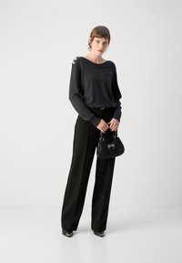 Pull en maille noir avec des boutons aux épaules, assorti à un pantalon noir à jambes larges. Tenant un petit sac à main noir, portant des chaussures à talons.