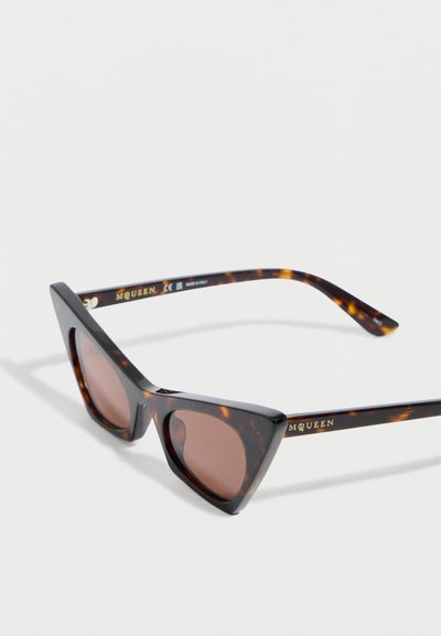 Γυαλιά ηλίου σε σχήμα γάτας με κεραμιδί σκελετό και καφέ φακούς, με το λογότυπο "MCQUEEN" στο μπράτσο, τοποθετημένα σε λευκή επιφάνεια.