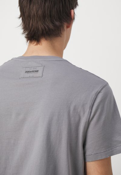 T-shirt in cotone grigio con collo girocollo, dettagli di cucitura visibili e un'etichetta in tessuto con la scritta "ZADIG & VOLTAIRE" vicino al collo.