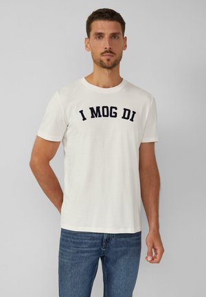 White cotton t-shirt with a crew neck and short sleeves, featuring navy blue text "I MOG DI" across the chest. Paired with blue jeans.