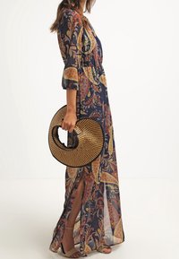 Lång, marinblå paisleymönstrad klänning med trekvartsärmar, genomskinligt tyg och sidofickor; håller en rund, vävd stroppväska.