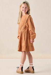 Robe orange-brun froncée avec des manches bouffantes et une broderie subtile. Possède une taille cintrée. Une paire de chaussures roses avec des accents noirs et des chaussettes rayées.