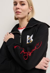 Crna zip-up dukserica s izvezenim crtanim logotipom patke i crvenim kurivim tekstom na rukavu. Mekani materijal sa opuštenim krojem.