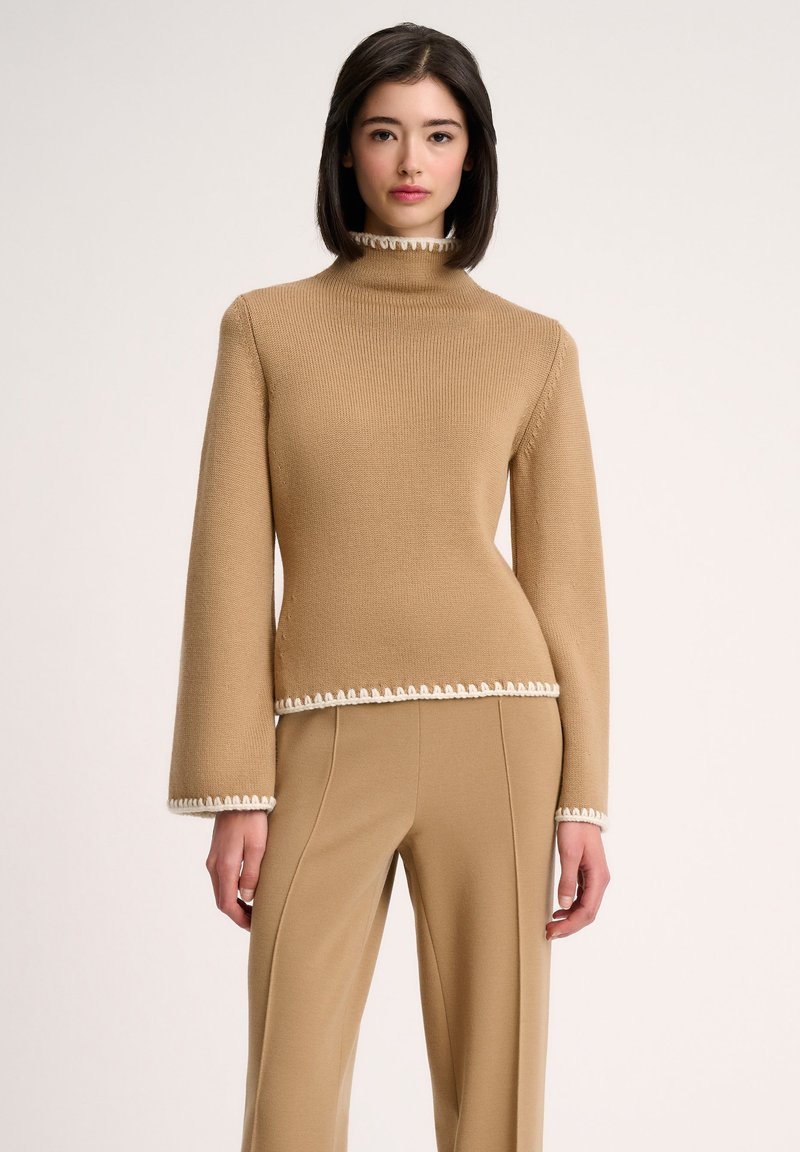 Pull en maille beige à col roulé avec des manches longues, présentant un motif texturé et une bordure festonnée blanche à l'ourlet et aux poignets.