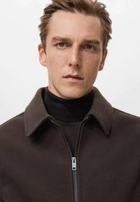 Ciemnobrązowa kurtka zapinana na suwak z kołnierzem, noszona na czarnym golfie. Gładka faktura, minimalne detale oraz srebrny zamek błyskawiczny jako akcent.