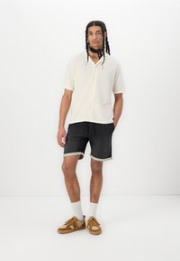 Chemise à boutons crème à manches courtes avec une finition texturée, associée à un short en denim noir avec des ourlets retroussés et des baskets en daim marron.