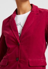 Henkilö, jolla on syvän punainen samettitakki valkoisen paidan päällä, jossa on musta abstrakti kuvio, näkyy kaulasta vyötärölle.