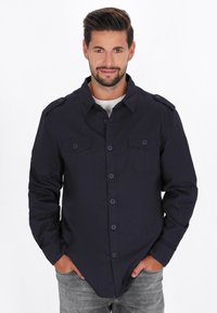 Marineblaues Hemd mit Knöpfen, doppelten Brusttaschen, strukturierten Schultern und Kragen. Der Stoff wirkt glatt und hat eine taillierte Passform.