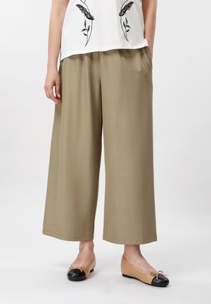 Žena má na sebe béžové krátke nohavice so širokými nohavicami, béžové a čierne baleríny s krytou špičkou a biely top s čiernou kvetinovou výšivkou.