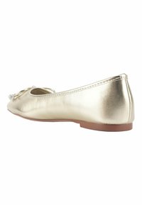 Scarpa piatta in oro metallizzato con punta arrotondata, texture liscia e un accento decorativo a fiocco. La suola è di un colore marrone chiaro.