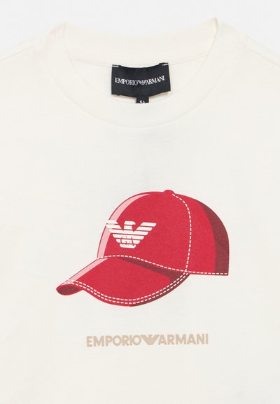 Λευκό πουκάμισο με γραφικό κόκκινο καπέλο μπέιζμπολ που φέρει το λογότυπο αετού της Emporio Armani και το όνομα της μάρκας κάτω από το καπέλο.