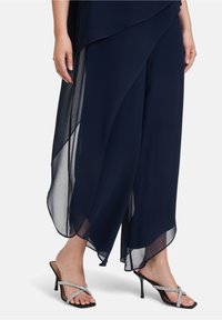 Granatowe, zwiewne spodnie z przezroczystym materiałem, z delikatnymi fałdami. W połączeniu z czarnymi sandałami na obcasie ozdobionymi srebrnymi akcentami.