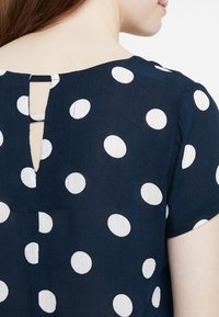 Mörkblå blus med vita prickar, med ett nyckelhål i nacken och korta ärmar, tillverkad av lätt texturerad tyg.