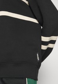 Sweat-shirt noir avec des rayures horizontales blanches sur la poitrine et les manches, ourlet côtelé et petite étiquette de marque ellesse sur la couture latérale.
