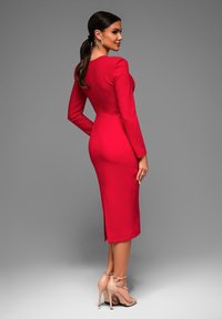 Gaun merah yang pas di badan dengan lengan panjang dan desain sepanjang lutut. Memiliki belahan belakang untuk kenyamanan bergerak. Dipadukan dengan sandal hak tinggi berwarna nude.