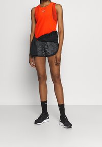 Czerwona bezrękawnik sportowy, czarne szorty z delikatnym wzorem, czarne buty do biegania i czarne skarpetki z logo; pozująca postawa.