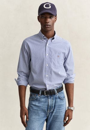 Homme portant une chemise à rayures bleues et blanches avec les manches retroussées, un jean bleu, une ceinture noire, une montre argentée et une casquette bleu marine avec la lettre G.