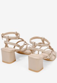 Sandalias de tacón cuadrado beige con tiras entrelazadas y hebillas en el tobillo. Material sintético liso con un ligero brillo y un robusto tacón cuadrado.