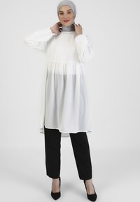 Bílá tunika s nařaseným pasem, dlouhými rukávy a splývavou, průsvitnou spodní vrstvou. V kombinaci s černými kalhotami a podpatky. Šedý hijab.
