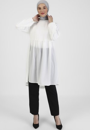 White tunic with a gathered waist, long sleeves, and a flowing, sheer lower layer. Paired with black trousers and heels. Gray hijab.