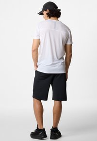 Biele športové tričko s krátkym rukávom a jemným švom v kombinácii s čiernymi športovými šortkami a čiernymi teniskami na jednoduchom pozadí.