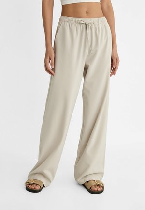 Kvinde iført løse beige bukser med snøre og beige sandaler, stående mod hvid baggrund, midterparti synligt.