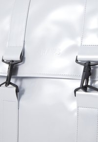 Weißer glänzender Rucksack mit schwarzem Zubehör, ausgestattet mit verstellbaren Trägern und geprägtem "RAINS"-Logo. Glatte Textur, minimalistisches Design.