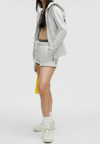 Femme portant un sweat à capuche gris clair, un short assorti roulé, un soutien-gorge de sport vert, des chaussettes blanches, des baskets blanches, tenant une bouteille d'eau jaune sur un fond uni.