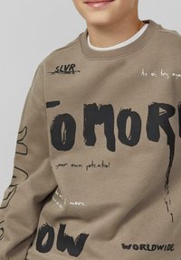 Bruine sweatshirt met zwarte grafische tekst en accenten. Heeft een ronde hals en lange mouwen met verschillende bedrukte zinnen en ontwerpen.