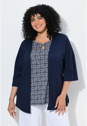 Smilende kvinde med krøllet hår iført en navy åben cardigan over en mønstret sort-hvid top og hvide bukser mod en ensfarvet baggrund.