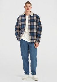 Young man standing wearing a blue and beige plaid jacket with shearling collar, white T-shirt, blue pants, and white sneakers against white background.