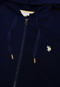 Sötétkék cipzáros kapucnis pulóver puha anyagból, ezüst logóval a mellrészen, texturált húzózsinórral és sima cipzáras záródással.