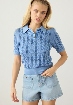 Young woman wearing a light blue cable knit short-sleeve polo sweater and high-waisted light denim shorts, standing against a plain background.