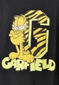 T-shirt noir avec un chat en dessin animé jaune, les bras croisés, sur un fond comportant une grande lettre 'G' jaune et des rayures de tigre. Texte "GARFIELD" en dessous.