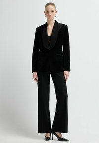 Zwarte fluwelen blazer met satijnen revers, voorzien van een borstzakje en twee voorzakken; gecombineerd met zwarte wijd uitlopende broek en puntige hakken.