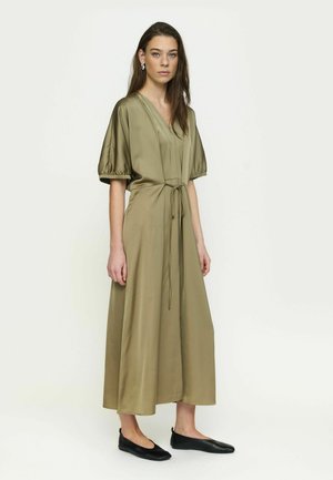 Moteris su ilgais rudos spalvos plaukais, dėvinti alyvuogių žalią midi suknelę su pūstomis rankovėmis ir juodus balerinas, stovinti prie paprasto šviesaus fono.