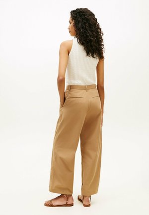 Femme aux cheveux bouclés, de profil, portant un haut en maille blanc sans manches, un pantalon beige à jambes larges et des sandales plates marron, debout les mains dans les poches.