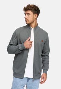 Hombre joven vestido con una chaqueta gris con cremallera sobre una camiseta blanca y jeans azul claros, mirando hacia su izquierda contra un fondo sencillo.