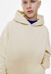 Beige huvtröja i mjukt material, med kengurufickor och liten logotyp på bröstet, bärs över en blå skjorta.