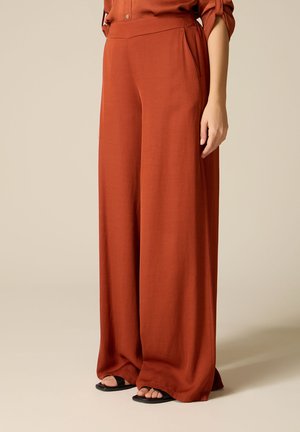 Pantaloni a gamba larga color ruggine con tasche laterali, indossati con sandali neri con punta aperta e top abbottonato ruggine coordinato.