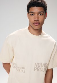 Beige Kurzarm-T-Shirt aus Baumwolle mit großem Text auf der Vorderseite. Lockerer Schnitt mit Rundhalsausschnitt.