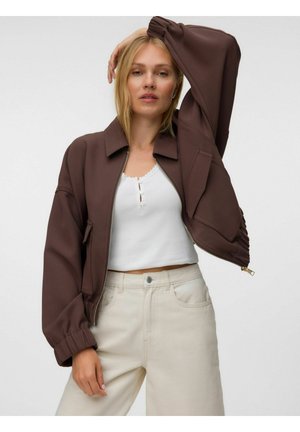 Femme aux cheveux blonds porte une veste zippée marron sur un top court blanc et un pantalon beige taille haute, posant avec un bras levé.