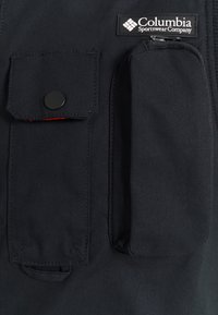 Black fabric vest featuring a snap-button chest pocket and a zippered side pocket, with an embroidered logo on the upper left.