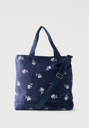 Marineblå stofnettaske med hvid blomsterbroderi, to hanke og en justerbar skulderrem på en ensfarvet baggrund.