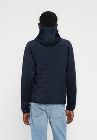 Άνδρας που φοράει σκούρο μπλε μπουφάν με κουκούλα και ραμμένες ραφές, συνδυασμένο με ανοιχτό μπλε τζιν, στέκεται στραμμένος προς τα πίσω μπροστά σε απλό φόντο.