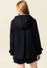 Cappotto nero con cappuccio realizzato in tessuto strutturato. Caratterizzato da una piega sul retro e maniche ampie con polsini. Indossato sopra pantaloni corti blu navy.